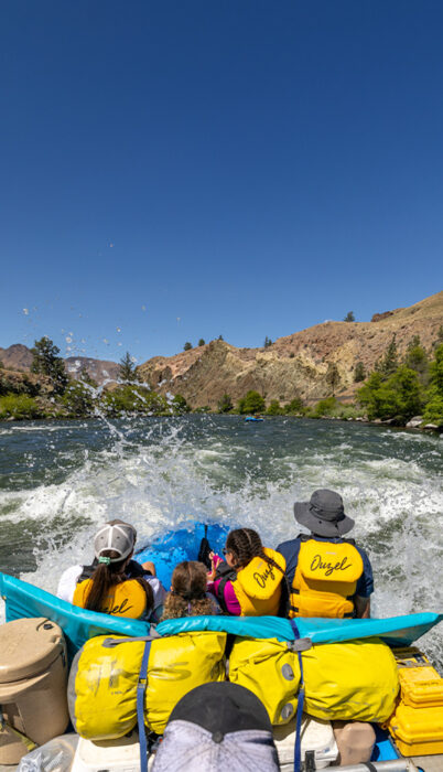 View from inside a raft as a family in yellow life jackets hits a splashy wave on the Deschutes River, surrounded by rocky hills and a bright blue sky.