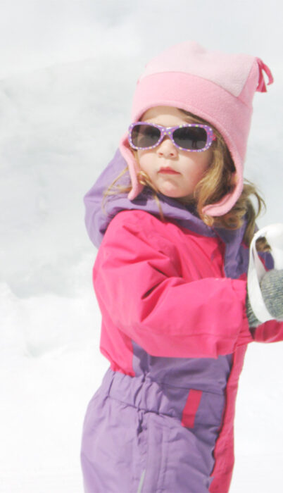Young girl in a pink snowsuit and sunglasses holds ski poles while enjoying a sunny winter day in Bend, Oregon.