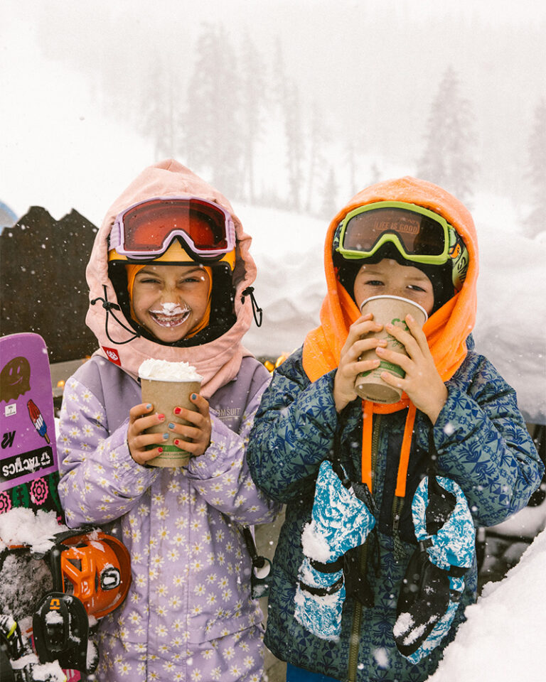 Two kids in colorful winter gear sip hot chocolate at Mt. Bachelor on a snowy day, smiling with whipped cream mustaches after a day of skiing in Bend, Oregon.