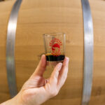 A close-up of a hand holding a small glass of dark beer from Deschutes Brewery in Bend, Oregon. The glass is lifted in front of a large wooden barrel inside the brewery. Perfectly captures the craft beer culture of the Bend Ale Trail and the must-try beers featured in this Bend craft beer guide.
