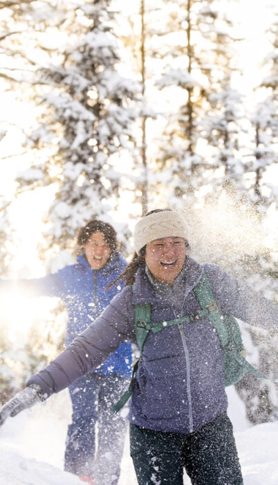 Two friends snowshoeing through fresh powder at Meissner Sno-Park near Bend, Oregon, laughing as sunlight filters through snowy trees—capturing the fun, affordable winter adventure Bend is known for.