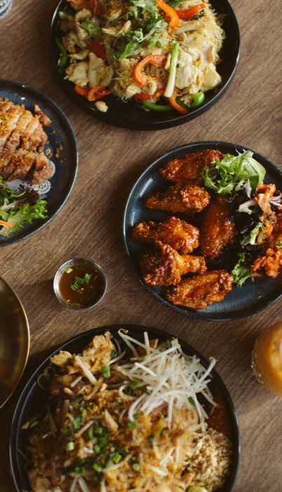 Overhead view of Thai-inspired noodle dishes, crispy chicken wings, and shared plates at a Bend, Oregon restaurant, showcasing some of the best food in Bend.