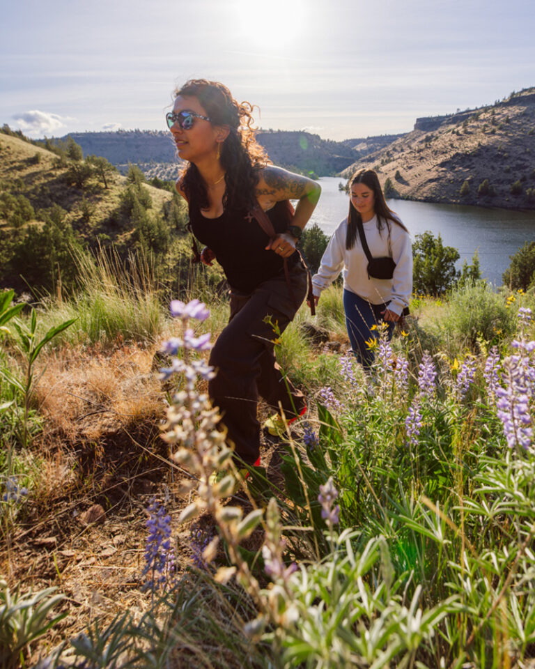 Two women hiking through spring wildflowers including purple lupine along the shores of Lake Simtustus near Bend, Oregon, with canyon walls and the Deschutes River in the background