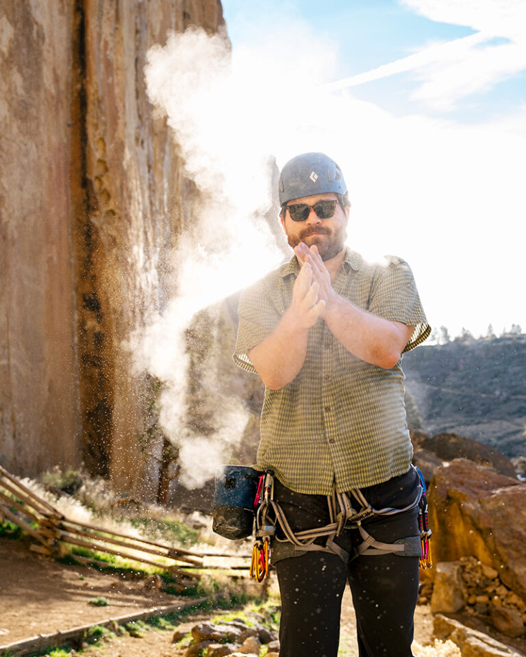 Male rock climber clapping chalk at Smith Rock State Park in Bend, Oregon, wearing a Black Diamond helmet and harness with the park's iconic volcanic tuff walls in the background