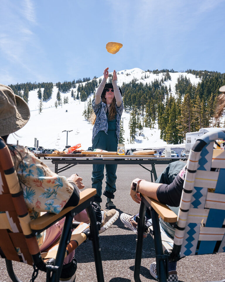 Group of friends enjoying a spring tailgate lunch at Mt. Bachelor ski resort in Bend, Oregon, with a woman tossing pizza dough in the air against a backdrop of snow-covered slopes and ski lifts.