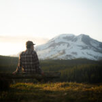 A man in a flannel shirt and cap sits on a log at golden hour along the Cascade Lakes Highway, looking toward a snow-covered South Sister rising above the tree line in the warm evening light.