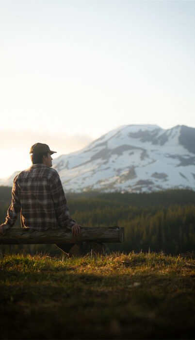 A man in a flannel shirt and cap sits on a log at golden hour along the Cascade Lakes Highway, looking toward a snow-covered South Sister rising above the tree line in the warm evening light.