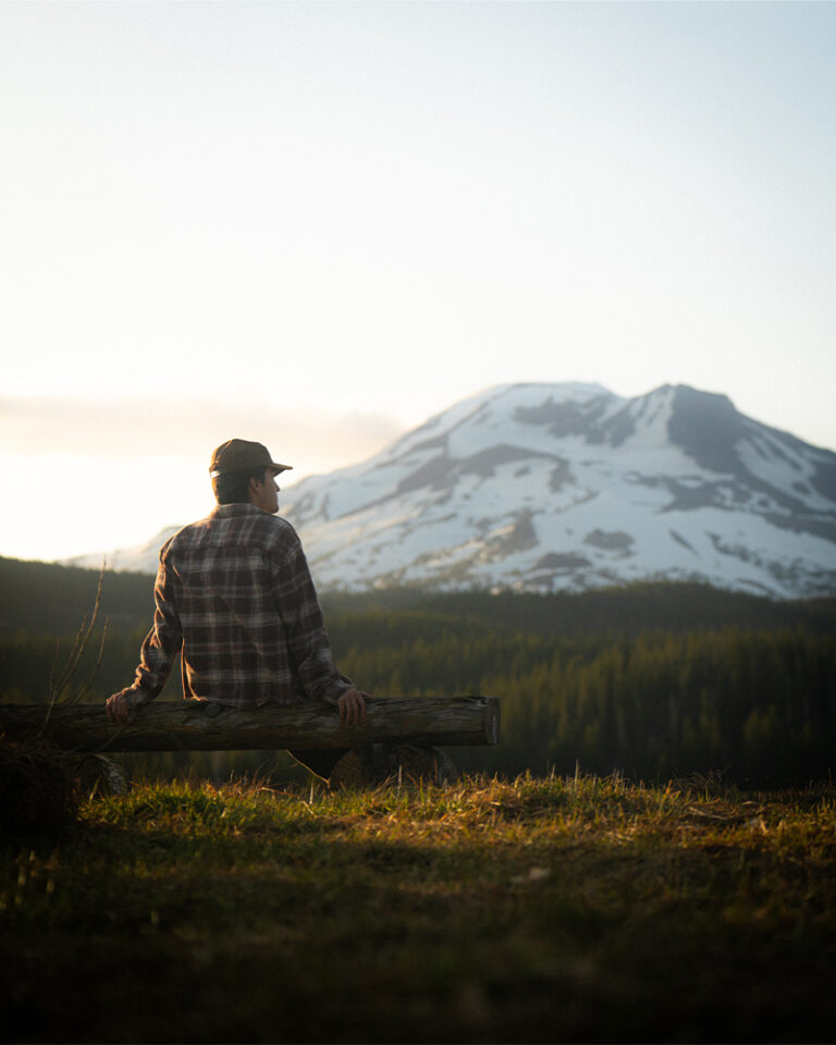 A man in a flannel shirt and cap sits on a log at golden hour along the Cascade Lakes Highway, looking toward a snow-covered South Sister rising above the tree line in the warm evening light.