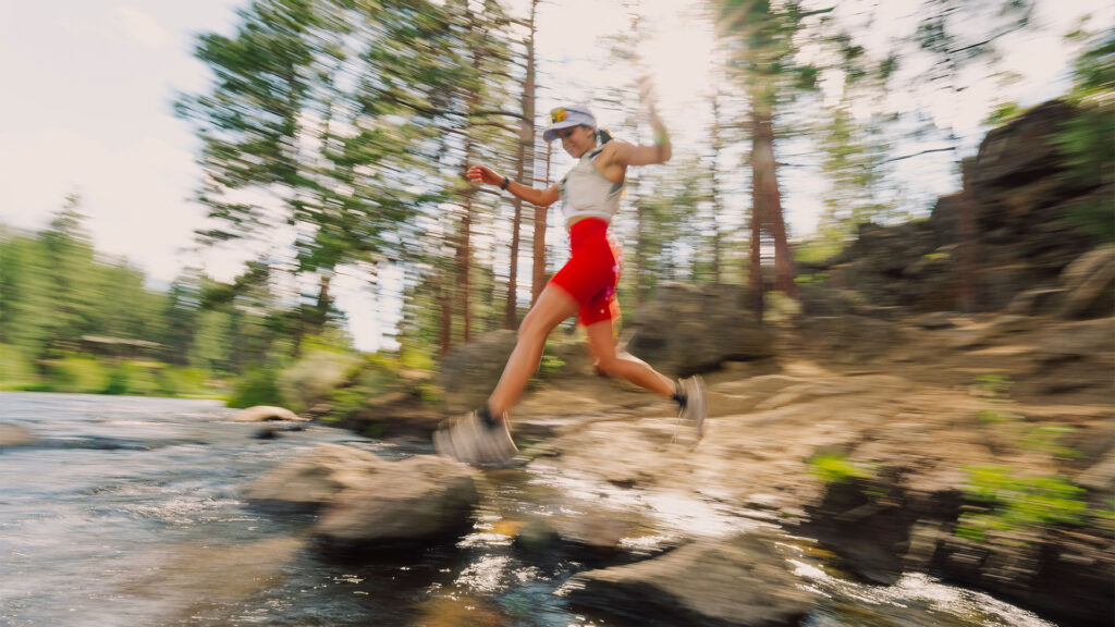 A trail runner leaps across rocky river stones in a sun-drenched forest, captured with motion blur to convey speed and energy, wearing red shorts and a light gray top.