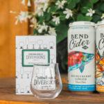 Drinkable Diversions passport booklet and tasting glass beside Bend Cider Co. cans, part of Bend's self-guided wine and spirit tasting trail