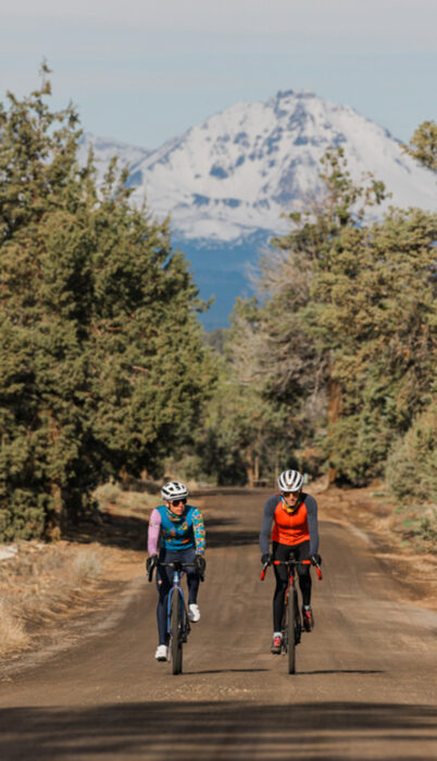 Two cyclists riding a gravel road near Sisters, Oregon with snow-capped Cascade mountains in the background.