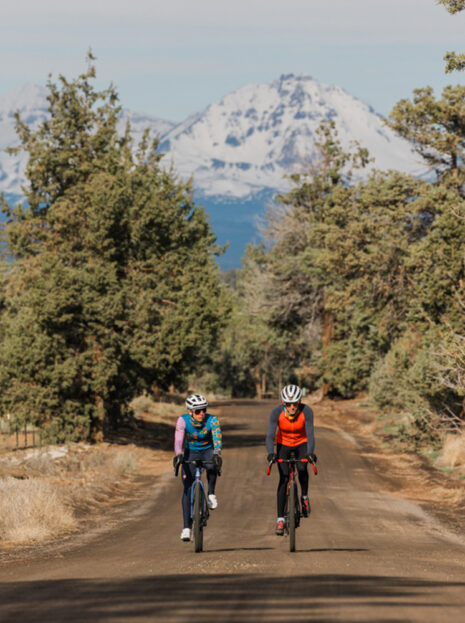 Two cyclists riding a gravel road near Sisters, Oregon with snow-capped Cascade mountains in the background.